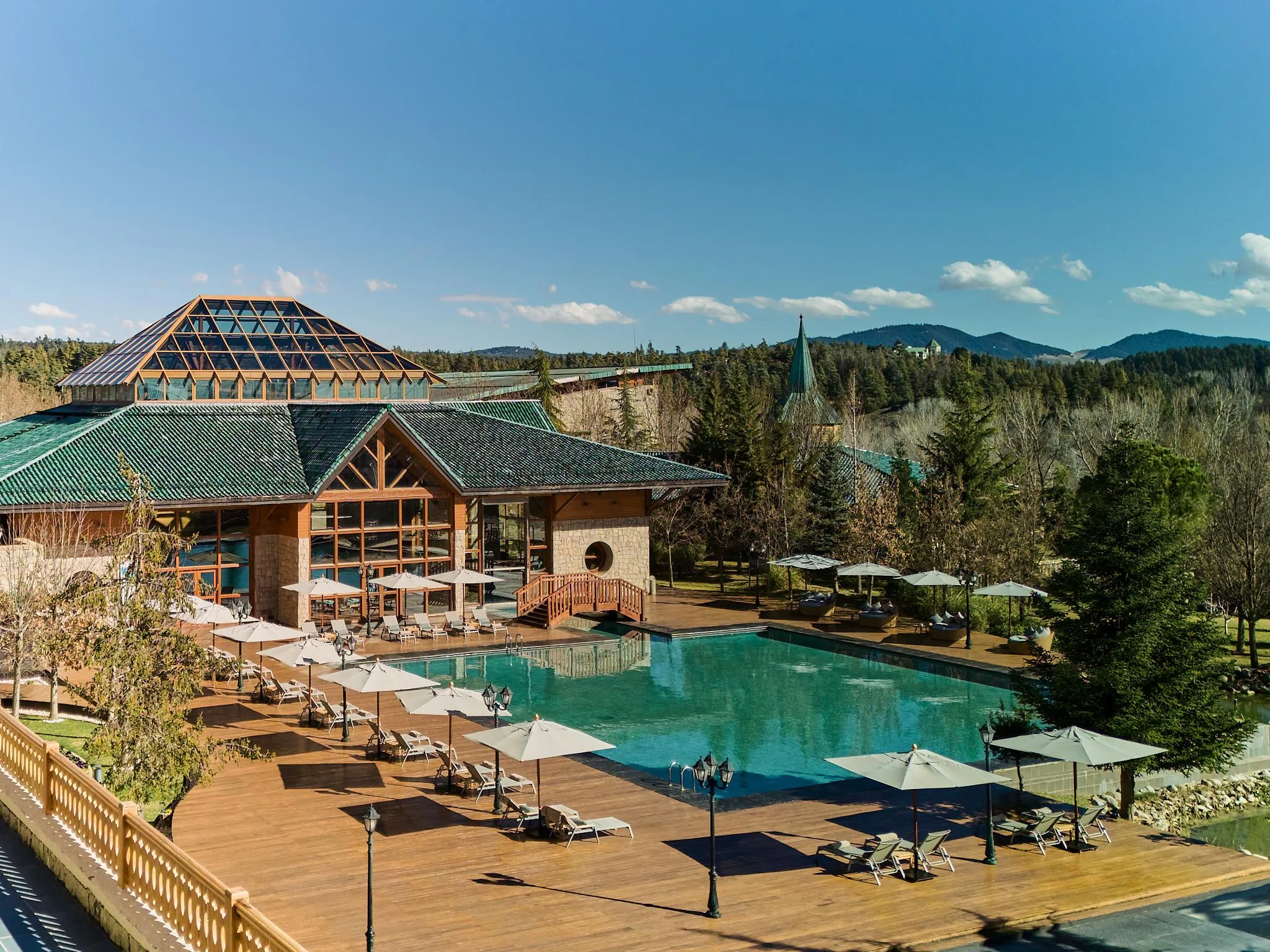 Piscine extérieure entourée de transats et de parasols, avec un bâtiment en bois et toit vert en arrière-plan, dans un cadre naturel montagneux