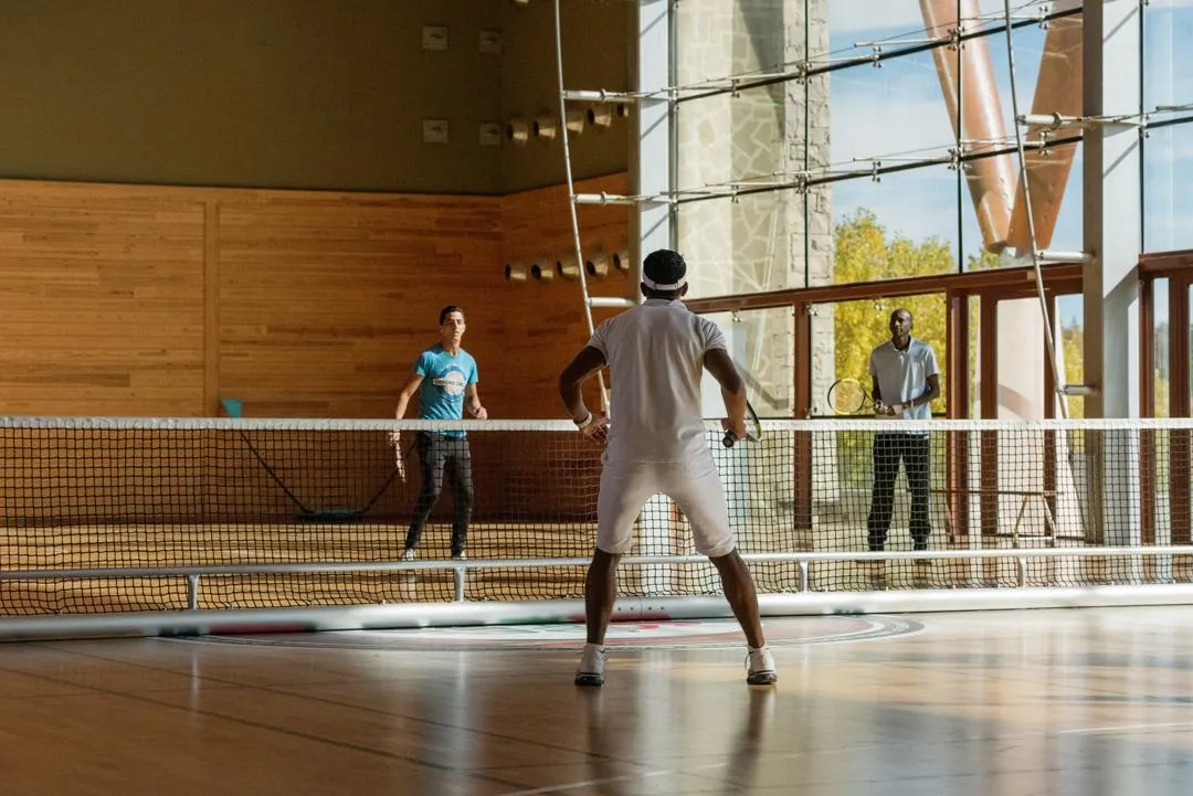 Trois personnes jouent au tennis de table dans une salle avec des murs en bois et de grandes fenêtres