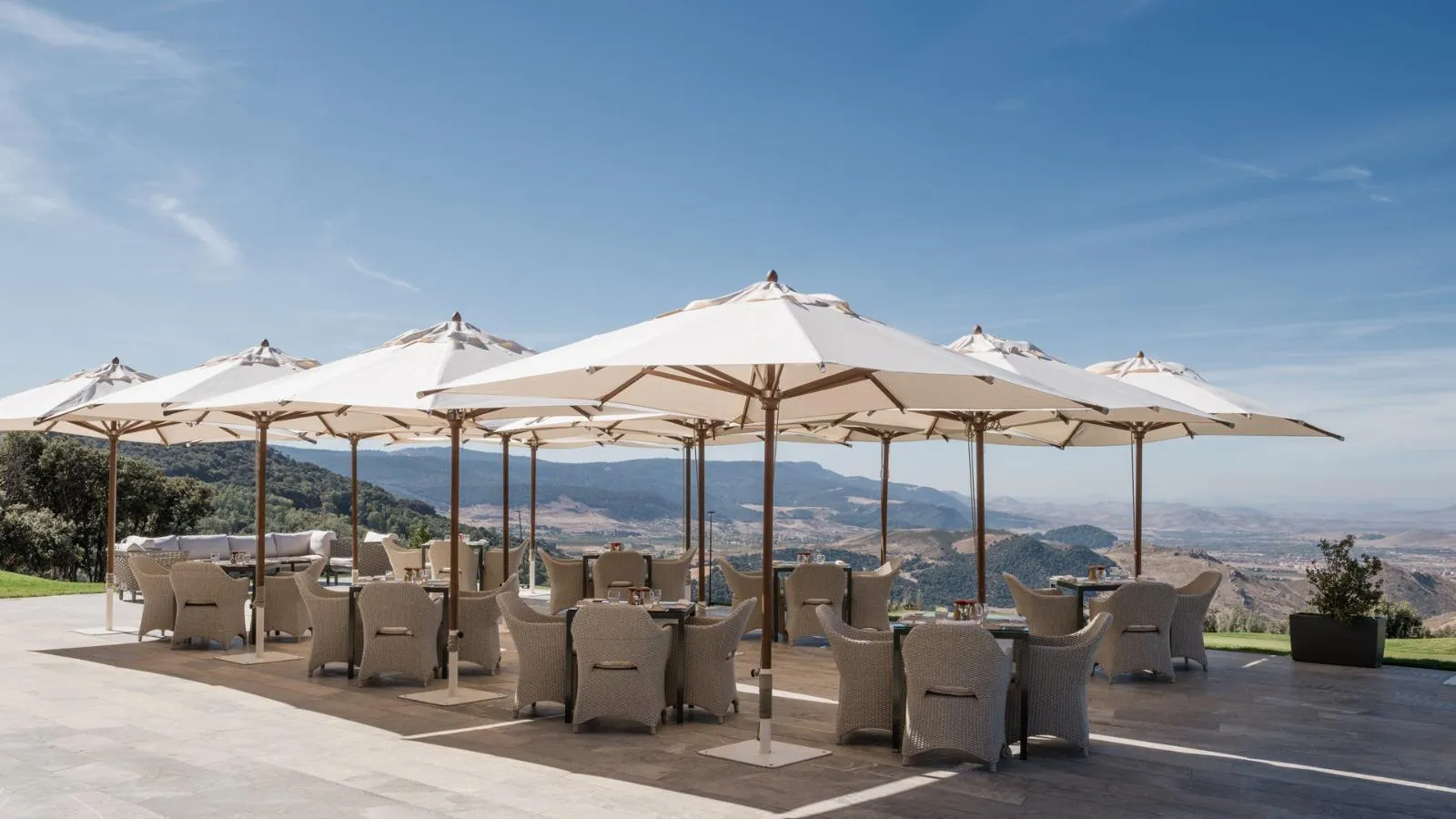 An outdoor terrace with tables, wicker chairs, and large umbrellas under a blue sky, offering a view of hills and mountains.