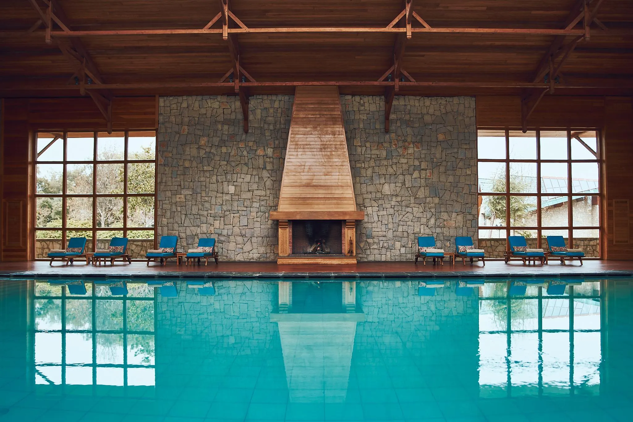 Piscine intérieure avec des chaises longues devant un mur en pierre et une cheminée allumée, entourée de grandes fenêtres en bois