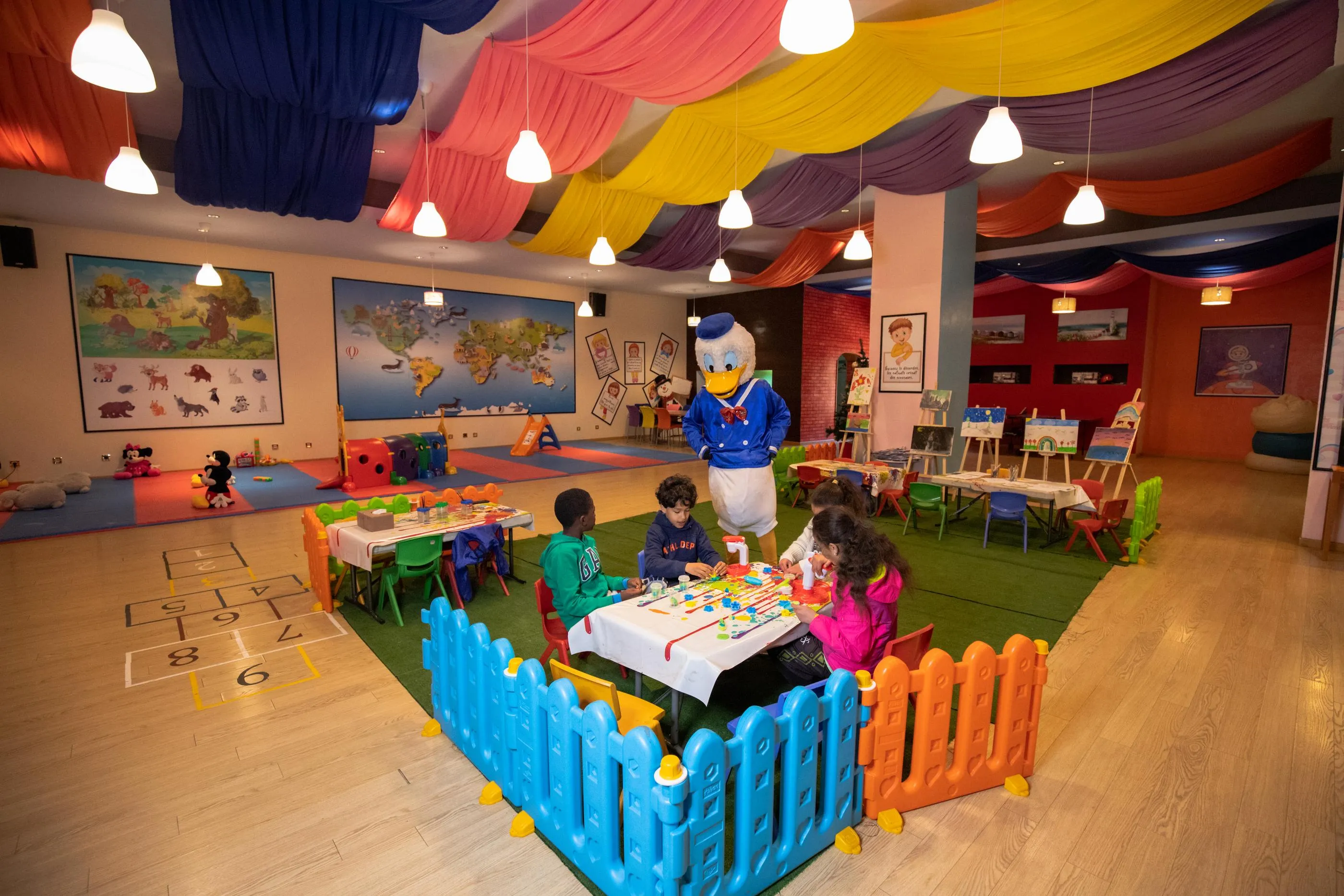 Une salle de jeux colorée avec des enfants assis à des tables, un personnage de dessin animé en costume et des décorations murales éducatives