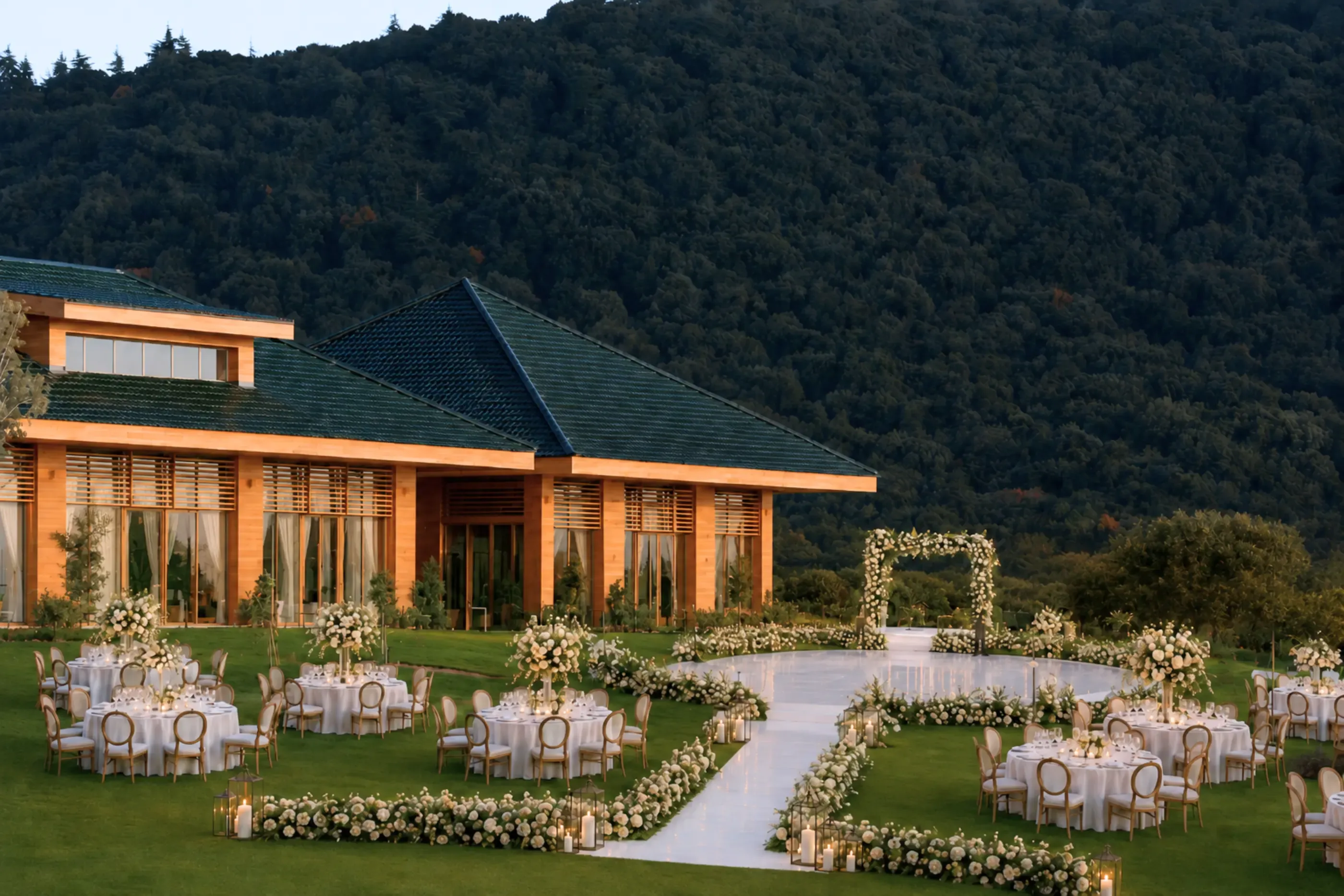 Un espace extérieur aménagé pour un événement avec des tables, des chaises et des décorations florales, devant un bâtiment aux murs orange et toit vert, entouré d'une colline boisée