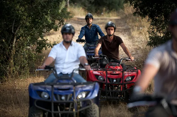 Trois personnes portant des casques roulent sur des quads dans un chemin de terre entouré de végétation