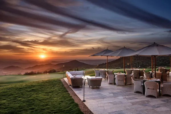 Terrasse avec des chaises et des tables sous des parasols, face à un coucher de soleil sur des montagnes et un terrain de golf