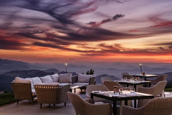 Terrace with wicker tables and chairs, white cushions and glasses, facing a sunset over mountains.