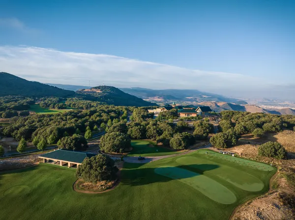 Vue aérienne d'un parcours de golf entouré d'arbres et de collines avec un bâtiment principal en arrière-plan