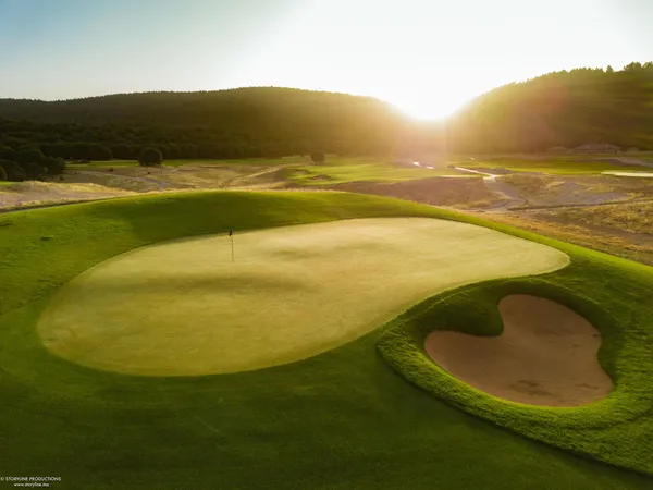 Un green de golf avec un drapeau au centre, entouré de bunkers de sable, sous un soleil couchant avec des collines boisées en arrière-plan
