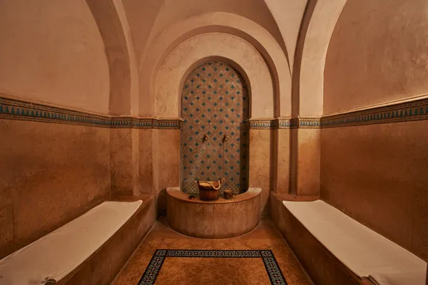 Salle de hammam avec des bancs en pierre, une niche carrelée de motifs géométriques et des seaux en métal