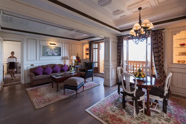 Salon avec canapé, fauteuils, table basse, table à manger et chandelier, une personne passe une porte avec un drap