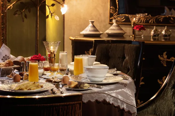Table dressée pour le petit-déjeuner avec des boissons, des œufs, des viennoiseries et des fruits, dans un cadre élégant avec des chandeliers et des coussins