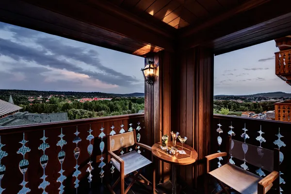 Balcon en bois avec table, chaises et verres, offrant une vue panoramique sur une ville et des collines sous un ciel nuageux