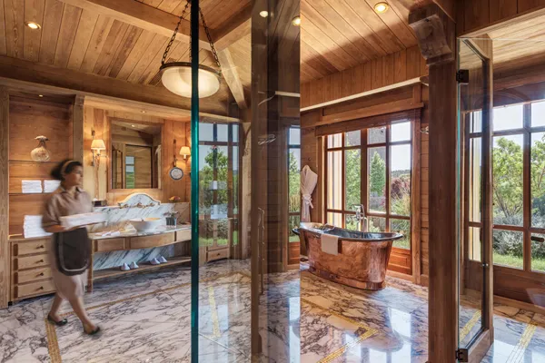 Une personne marche dans une salle de bain en bois avec une baignoire en cuivre, un lavabo en marbre et de grandes fenêtres donnant sur l'extérieur