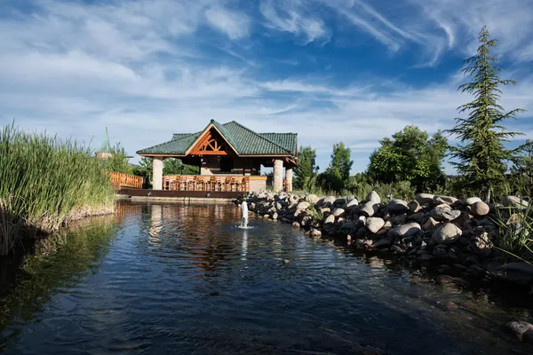 Un pavillon en bois avec un toit vert situé au bord d'un plan d'eau entouré de rochers et de végétation