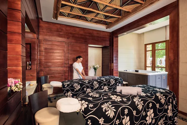 A person in white attire prepares a massage table in a wooden spa room with windows and plants.