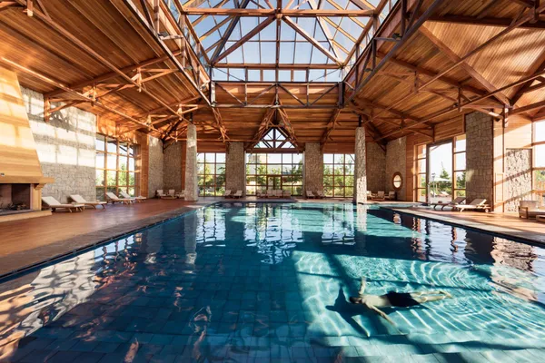 Piscine intérieure avec un toit en bois et des grandes fenêtres, entourée de chaises longues et de colonnes en pierre