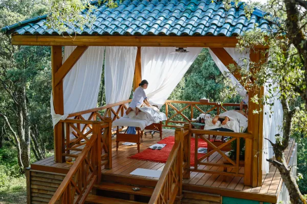 Deux personnes allongées sur des tables de massage en plein air sous un auvent en bois avec un toit en tuiles bleues, entourées d'arbres