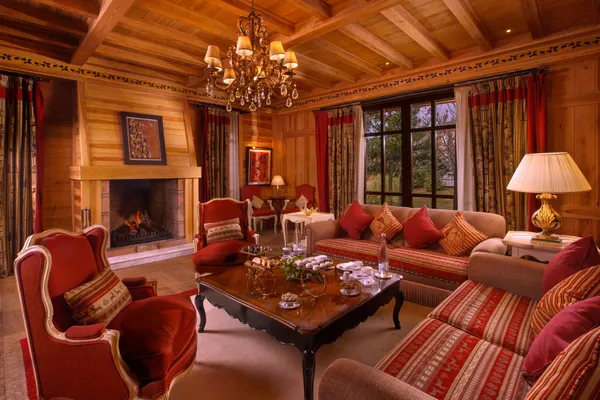 Salon en bois avec cheminée allumée, canapés et fauteuils rouges, table basse avec thé et chandeliers, lustre et grandes fenêtres